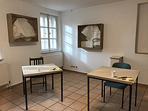 Der Kunstübungsraum. In der Mitte stehen zwei Werktische, an denen jeweils eine Person arbeiten kann. An den Wänden hängen in dreidimensionale Rähmen gefasste Plastiken aus Papier.