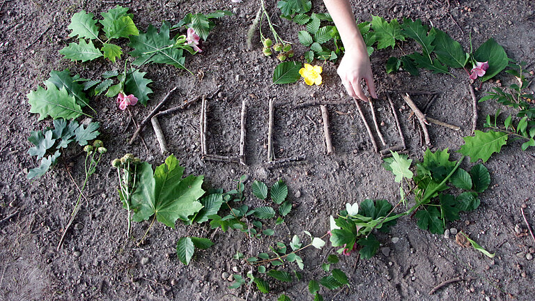 Auf Erdboden legt eine hand das Wort "Kultur" mit Stöcken. Außen herum bilden Baumblätter eine Herzform.  