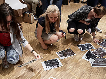 Auf dem Boden liegen schwarz-weiß Fotografien, davor sind drei Frauen in der Hocke und betrachten die Fotos oder halten welche in der Hand.