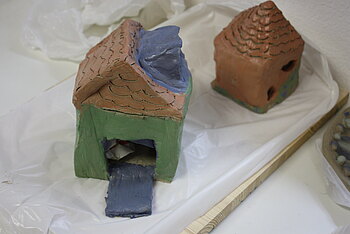 Zwei Modellhäuser aus gebrannter Keramik stehen auf einem weißen Tuch. Ein Haus ist ganz braun glasiert. Das andere Haus hat ein Braunes Dach, grüne Wände und einen blauen Kamin.