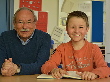 Ein älterer Mann und ein Junge im Grundschulalter sitzen an einem Tisch und lächeln. Vor dem Jungen liegt ein aufgeschlagenes Buch. Im Hintergrund ist ein Klassenzimmer zu sehen.