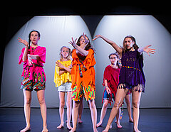 Foto zeigt Szene aus der Präsentation des Tanz LABs. Die Spielerinnen stehen in einer Formation vor einem weißen Hintergrund und tanzen gemeinsam eine Choreografie