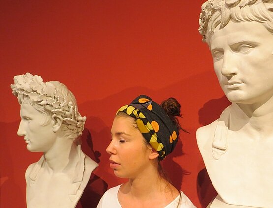 Der Kopf einer Frau ist zwischen zwei antiken Büsten vor einer roten Wand zu sehen.