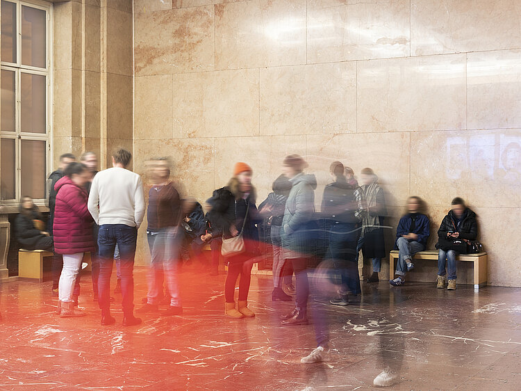 Eine Gruppe von Personen steht und geht in einem hohen Raum im Haus der Kunst. An den Wänden sitzen weitere Personen auf Bänken. Durch Langzeitbelichtung sind die Personen auf dem Foto nur verschwommen zu sehen.