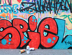 Das Wort "Spiel" ist in roter Schrift auf eine Wand gesprayt; davor stehen und liegen Graffiti-Spraydosen.