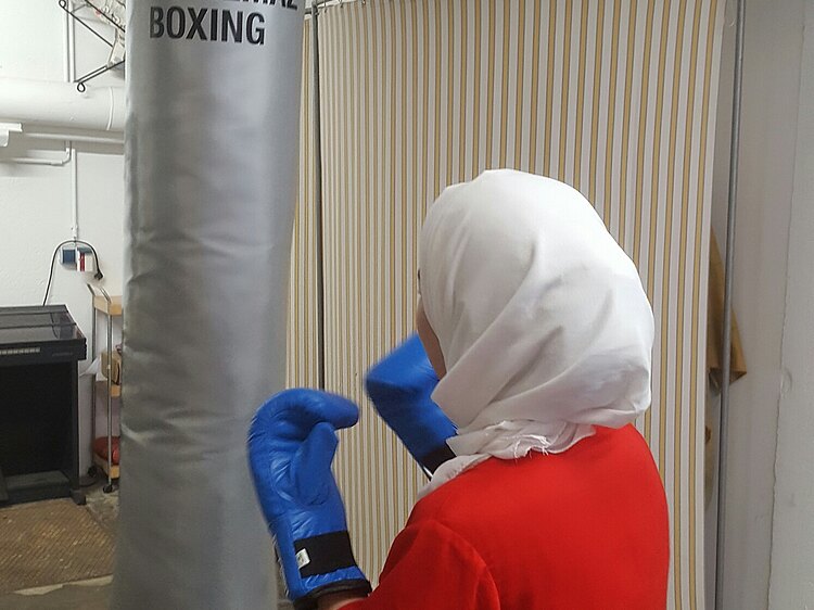 Frau mit Boxhandschuhen und Kopftuch vor Boxsack