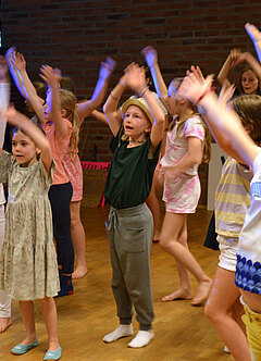 Eine große Gruppe von Kindern singt und tanzt mit erhobenen Händen in einem Probenraum.