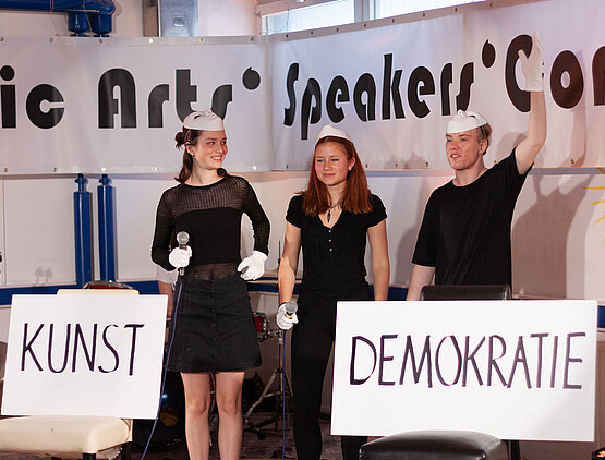 Drei Jugendliche auf Bühne mit Schildern “Kunst” und “Demokratie”