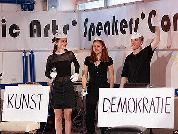 Drei Jugendliche auf Bühne mit Schildern “Kunst” und “Demokratie”