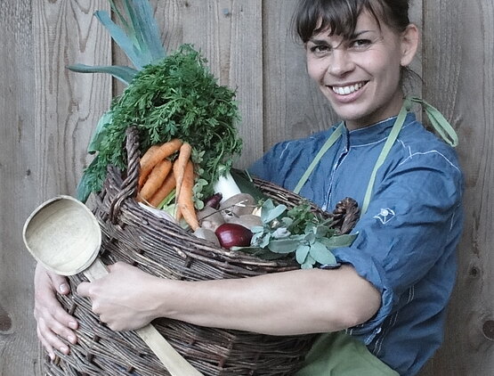 Eine Frau hält einen Korb voll mit Gemüse lächelnd in die Kamera