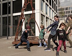Foto zeigt einen Moment aus einer Theaterprobe draußen in der Stadt. Eine Gruppe Kinder kopiert den Umriss einer Skulptur.