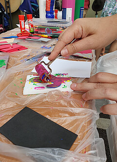Der Bildausschnitt zeigt die Hände einer Person, die mit einer Farbrolle Farbe auf einen Stempel aufträgt, mit dem danach eine Karte bedruckt werden wird.