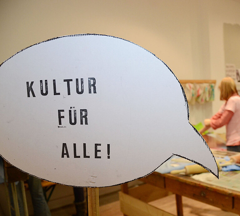 Der Text "Kultur für alle!" steht in einer Sprechblase, im Hintergrund sieht man Kinder in einer Buchwerkstatt.  