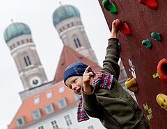 Kind hängt an Kletterwand; im Hintergrund ist der Liebfrauendom zu sehen