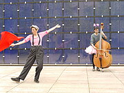 Der als Frau verkleidete Schauspieler spielt Cello während die Frau, welche als Mann verkleidet ist zu der Musik tanzt