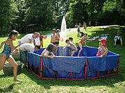In einem Park ist ein großes, mobiles Plantschbecken aufgebaut. Viele Kinder in Badekleidung spielen darin oder rennen darum herum.