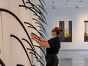 Eine junge Frau mit Dunkelbrille steht vor einer Ausstellungswand. Sie erstastet eine Objektgruppe mit aus der Wand herausragenden längliche und spitz zulaufenden Formen.