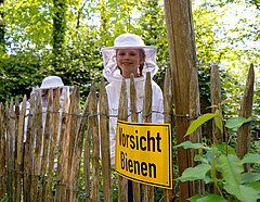 Zwei Kinder stehen mit Schutzanzügen hinter einem Zaun und am Zaun ist ein Schild mit der Aufschrift "Vorsicht Bienen"