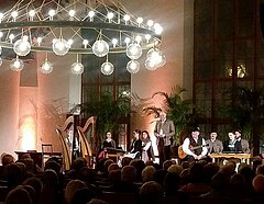 Harfenspieler und weitere Musiker stehen auf einer Bühne vor einem großen Publikum und tragen traditionelle bayrische Kleidung