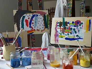 zwei Staffeleien mit Bildern, gemalt mit selbstgemachten Eitemperafarben. Auf dem Tisch sind Pigmente, die Ei-und-Leinölemulsion, Glasplatten und Pinsel zu sehen.