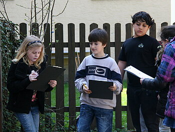 Kinder stehen vor einem Zaun mit Klemmbrettern in der Hand und machen sich Notizen