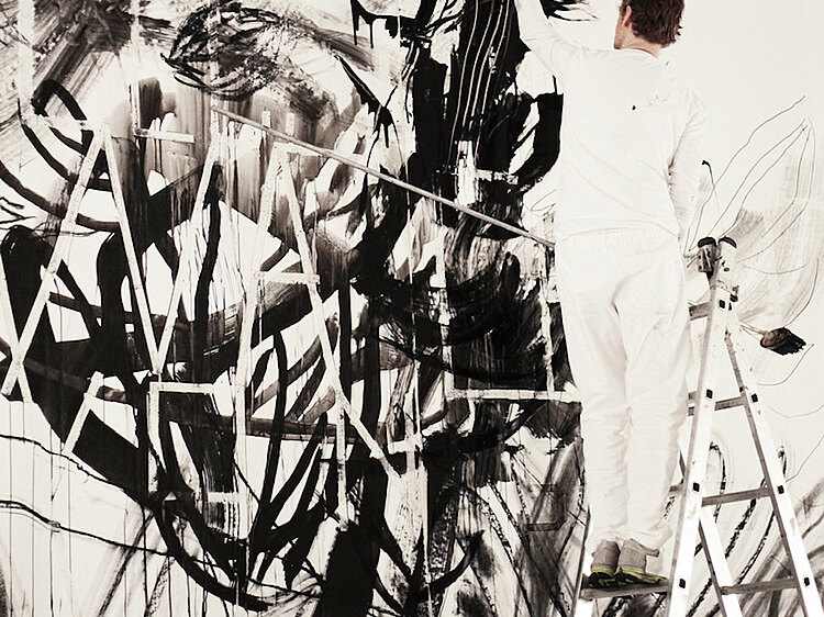 Ein Mann in weißer Kleidung steht auf einer Leiter und malt dicke schwarze Striche auf die Wand
