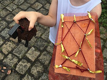 Eine Kinderhand hält ein braunes Holzschäfchen, die andere eine Holzplatte auf die mit Nägeln eine rote Schnur befestigt ist, ein rotes Obstnetz und goldene Stofffetzen