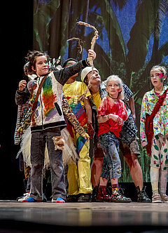 Acht bunt kostümierte Kinder blicken dem Pfeil hinterher, den ein im Vordergrund stehendes Kind mit Bogen in der Hand nach rechts geschossen hat. Es handelt sich um eine Bühnenszene aus „Peter Pan“.