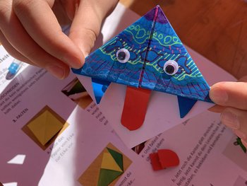 Ein Lesezeichen, das aus Origamipapier gefaltet wurde. Es ist mit Mustern, angeklebten Augen und einer Zunge verziert.