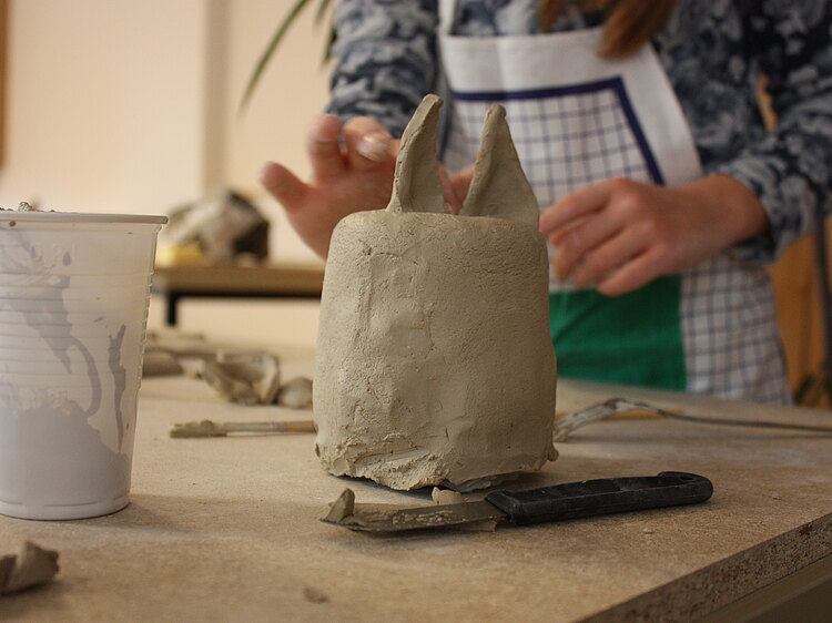 Detailaufnahme einer Keramik. Die Arbeit hat die Form eines Zylinders und oben sind zwei spitze Ohren befestigt. Die Keramik ist noch nicht gebrannt oder glasiert.