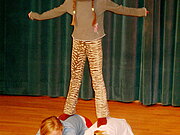 Drei Mädchen machen zusammen Akrobatik, ein Mädchen steht auf dem Rücken zwei anderer Mädchen während diese in embionaler Position auf dem Boden liegen