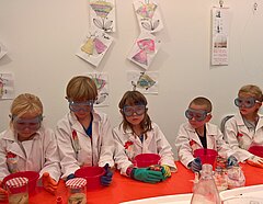 Fünf Kinder sitzen bereit für ein Experiment mit Brille und Schutzmantel an einen Tisch