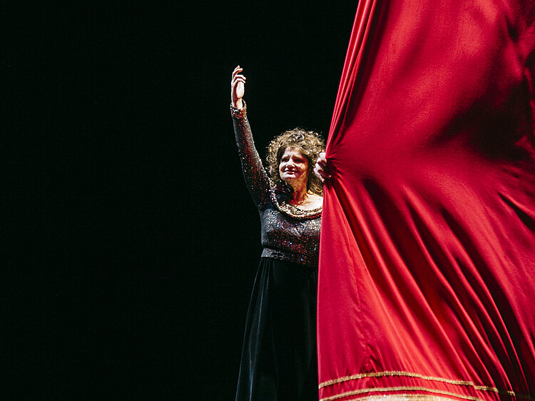 Eine Frau in schwarzem Kleid öffnet einen roten Vorhang