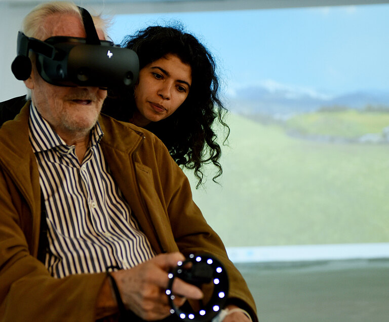 Eine Jugendliche steht hinter einem älteren Mann, der eine VR-Brille benutzt.  