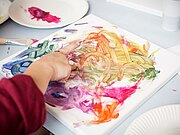 Eine Person malt mit Fingern und verschiedenen Farbe auf eine Leinwand.