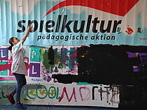Bemalte Wand mit Schriftzug "Spielkultur"; am linken Bildrand springt ein Kind in die Höhe.