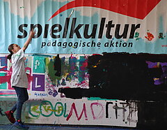 Bemalte Wand mit Schriftzug "Spielkultur"; am linken Bildrand springt ein Kind in die Höhe.