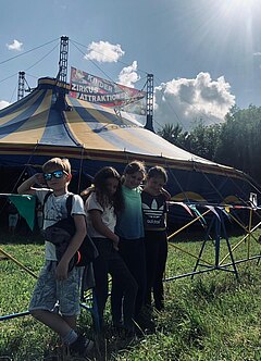 Vier Kinder lehnen am Zaun vor einem blau-gelb gestreiften Zirkuszelt. Über dem Zelt steht auf einem Schild "Kinder Zirkus Attraktionen".