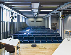 Blick in den Filmsaal des Pädagogisches Instituts: Mehrere Reihen blauer Kinostühle füllen den Raum. Im vorderen Bereich ist eine Holzbühne mit zwei Tischen, Mikrofonen und Stühlen für Redner*innen.