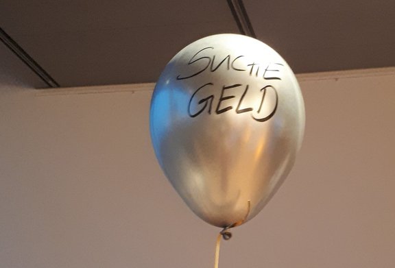 Auf einem silberfarbenen heliumgefüllten Luftballon, der an einer Schnur hängt, steht "Suche Geld".  