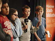 Eine Gruppe von Kindern steht auf der Bühne und spricht abwechselnd ins Mikrofon.