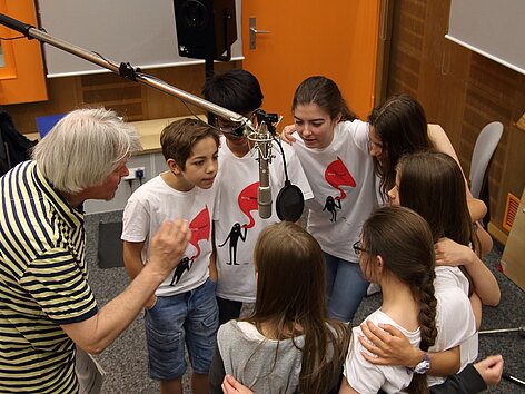Sieben Kinder und ein Stimmtrainer stehen um ein Mikrofon herum