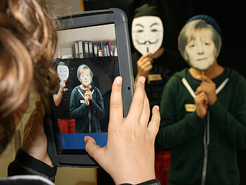 Ein Kind fotografiert zwei Kinder mit einer Angela Merkel Maske und einer Maske mit Schnurrbart
