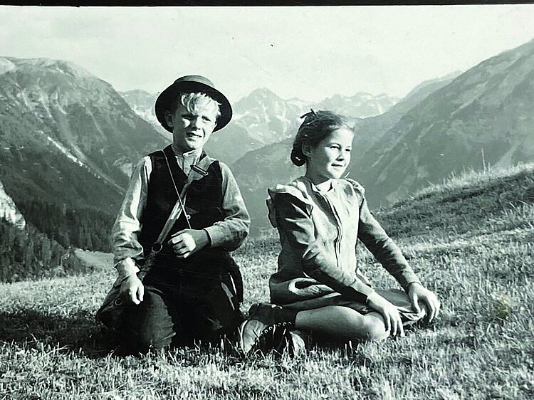 Heidi und Peter sitzen auf einer Wise vor Bergen.