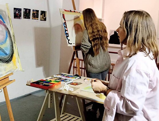 Zwei junge Frauen malen in einem Atelierraum an Staffeleien mit Kreide auf Papier. Zwischen ihnen steht ein Tisch mit Kreide.