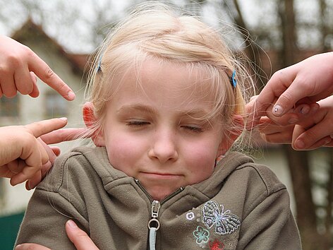 Ein Mädchen kneift ihre Augen zu und zieht ihre Schultern nach oben während fünf Hände versuchen ihr die Ohren zuzuhalten