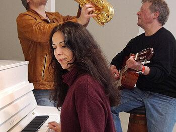 Eine Frau spielt Klavier während ein Mann Trompete spielt und ein weiterer Gitarre spielt