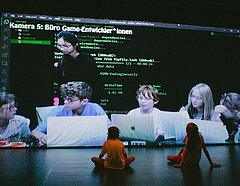 Foto zeigt eine Szene aus einer Präsentation eines Theaterkurses. Zwei Spieler*innen sitzen vor einer großen Videoprojektion mit dem Rücken zum Publikum. 