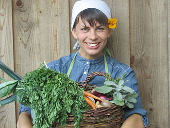 Eine Frau hält einen Korb voll mit Gemüse lächelnd in die Kamera