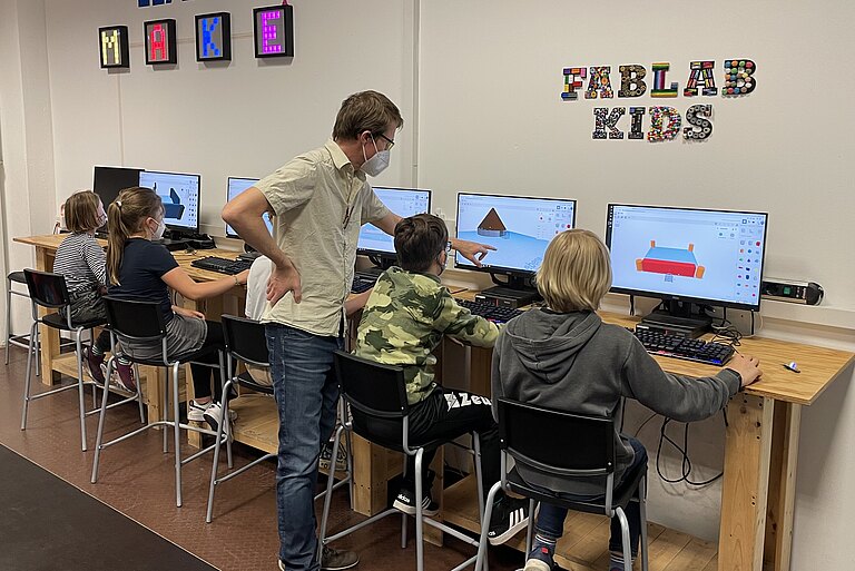 Fünf Schulkinder arbeiten an Computern. Ein Workshopleiter hilft dabei einem Kind.